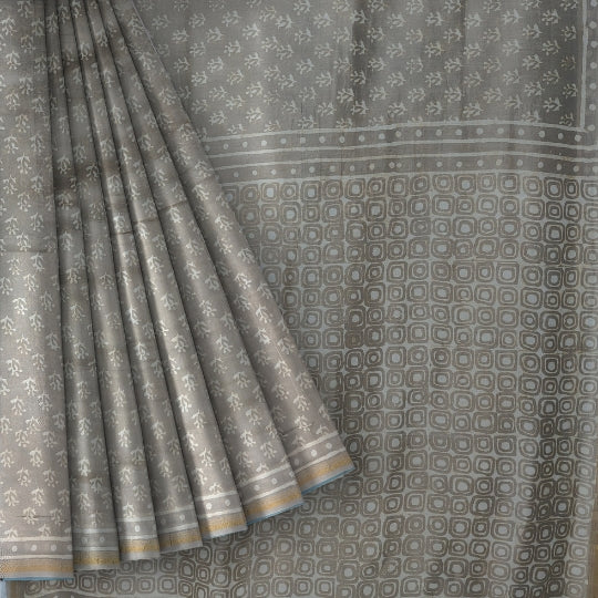 A close-up of a gray saree or fabric panel with a repeating small floral pattern and a border of alternating square and dot motifs, shown with one corner folded back to reveal the edge.
