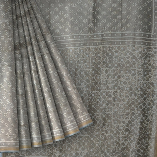 A close-up of a gray saree or fabric panel with a repeating small floral pattern and a border of alternating square and dot motifs, shown with one corner folded back to reveal the edge.