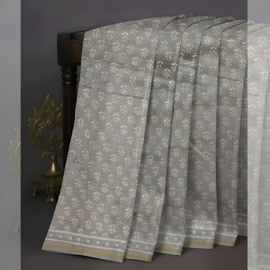 A light gray and white patterned saree draped elegantly over a dark wooden stand, featuring a decorative gold border and a traditional block print motif.