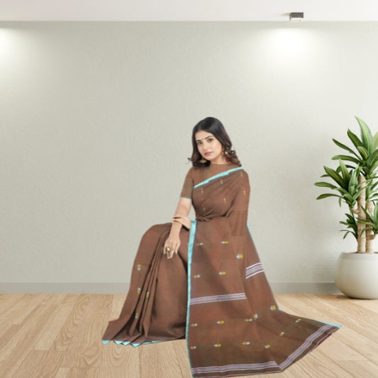 A woman wearing a brown saree with light blue border accents and small floral motifs, seated in a minimalist room with a wooden floor and a potted plant.