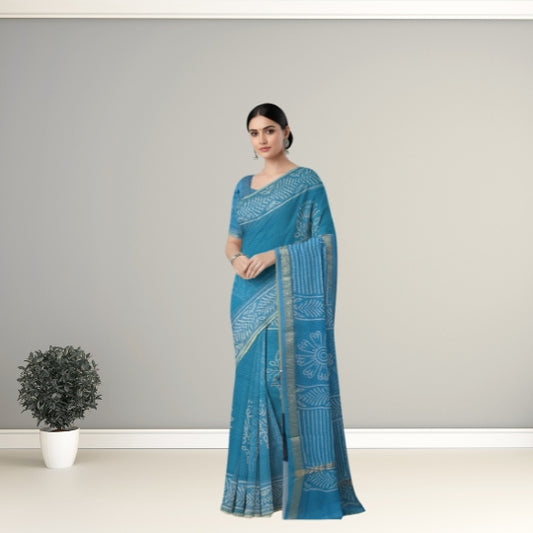 A woman modeling a teal blue saree with white block prints and a gold border, standing in a studio setting with a small potted plant on the left.