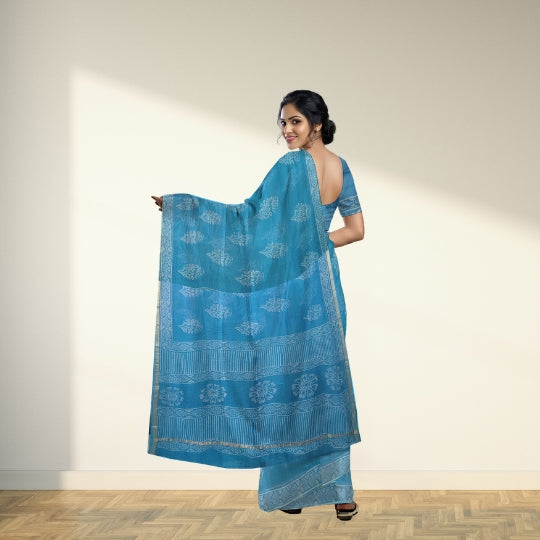 A woman modeling a blue saree with white block prints and a matching blue blouse, standing in a studio with a beige wall and wooden floor.