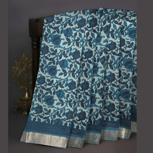 A draped blue and navy floral patterned fabric with a metallic silver border, displayed on a dark wooden stand next to a brass decorative object against a dark gray background.