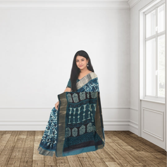 A woman wearing a blue and teal patterned saree with a metallic silver border, standing in a bright room with white walls and wooden floors.