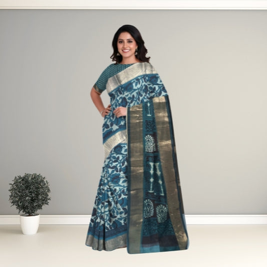 A woman modeling a blue and teal patterned saree with metallic gold borders, standing against a neutral gray studio background with a small potted plant on the left.