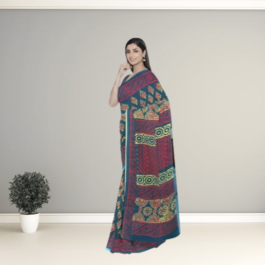 A woman modeling a dark teal blue saree with intricate red, green, and gold geometric patterns, standing in a minimalist studio setting with a small potted plant on the left.