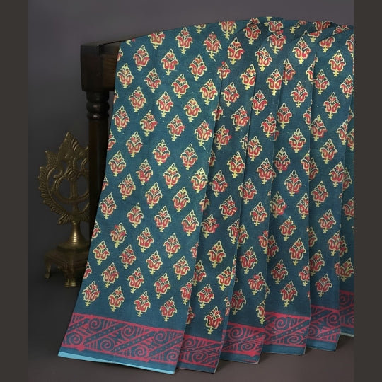 A dark teal blue saree with a repeating red and yellow floral block print and a red patterned border, draped over a dark wooden stand.