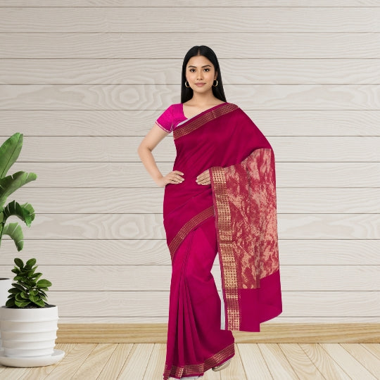 A woman modeling a magenta pink saree with a gold patterned border and a matching pink blouse, standing against a light wood paneled wall with a potted plant on the left.