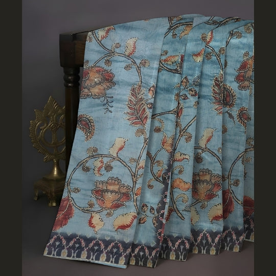 A light blue patterned fabric, likely a sari, draped over a dark wooden stand. The fabric features a detailed floral and vine motif in rust red, beige, and brown, with a decorative ikat-style border at the bottom.