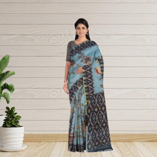 A woman modeling a blue and black patterned saree with a dark gray sleeveless blouse, standing against a light wood-paneled wall with a potted plant on the left.