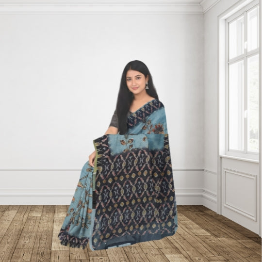 A woman modeling a blue and navy patterned saree with intricate gold and brown motifs, standing in a bright room with white walls and wooden floors.