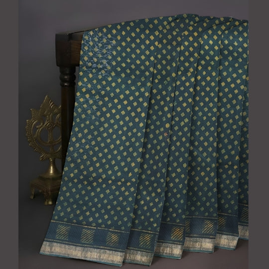 A dark teal blue saree with a repeating gold geometric pattern and a wide decorative border, draped over a dark wooden stand next to a brass decorative object.