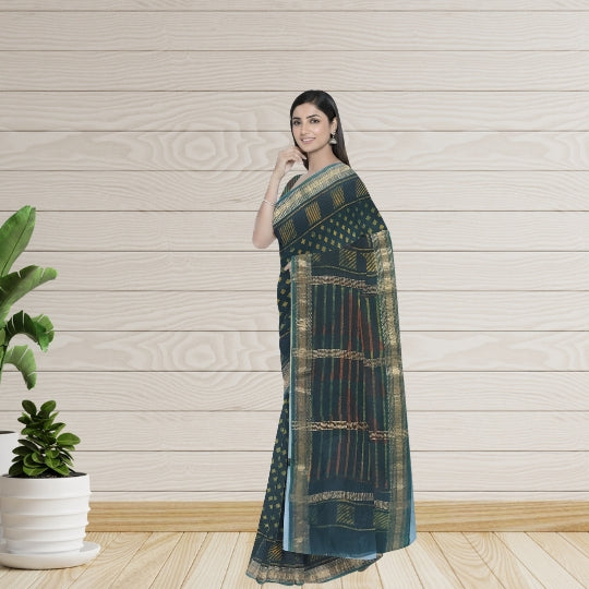 A woman modeling a dark teal-green saree with gold and red woven patterns, standing against a light wood-paneled wall with a potted plant on the left.