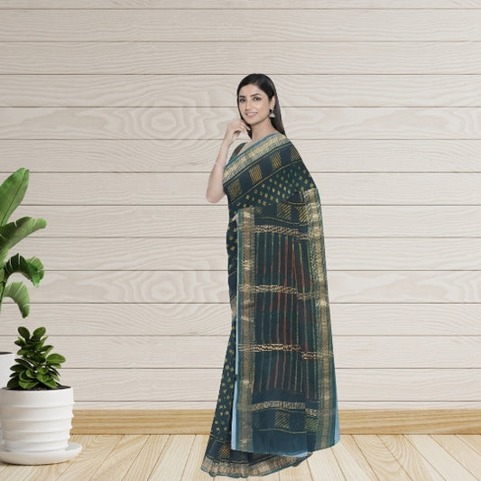 A woman modeling a dark teal-green saree with gold and red woven patterns, standing against a light wood-paneled wall with a potted plant on the left.