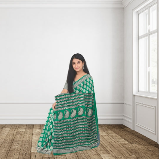 A woman wearing a green saree with white and silver patterns stands in a bright room with white walls and wooden floors, holding up the saree drape to show the design.