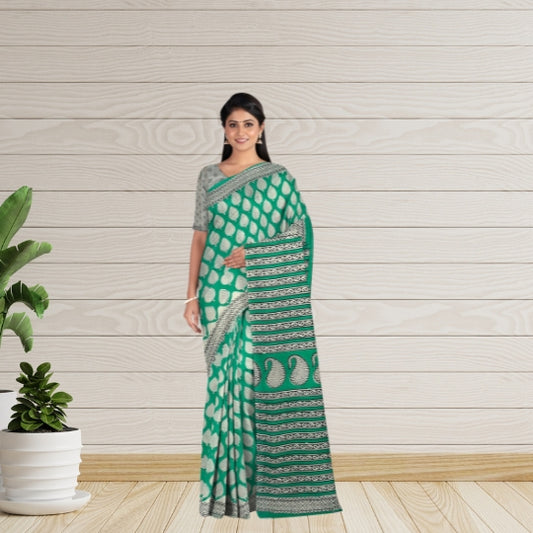 A woman modeling a green and white patterned saree with a matching printed blouse, standing against a light wood-paneled wall with a potted plant on the left.