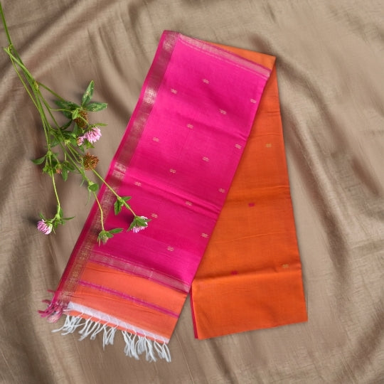 A folded bright pink and orange silk saree with white fringe and small metallic motifs, laid flat on a beige fabric background with a small sprig of green leaves and pink flowers.