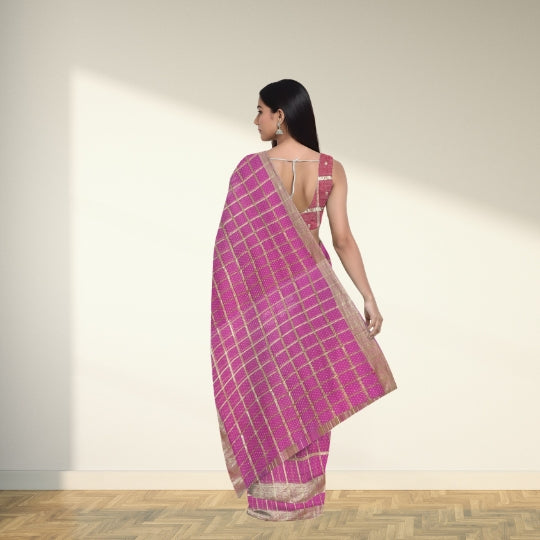 A woman wearing a bright pink saree with a gold grid pattern and a matching pink blouse, standing in profile against a warm beige wall with soft sunlight and a wooden floor.