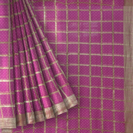 A close-up of a pink fabric, likely a saree, featuring a gold grid pattern and small gold dots, with a pleated drape showing the border.