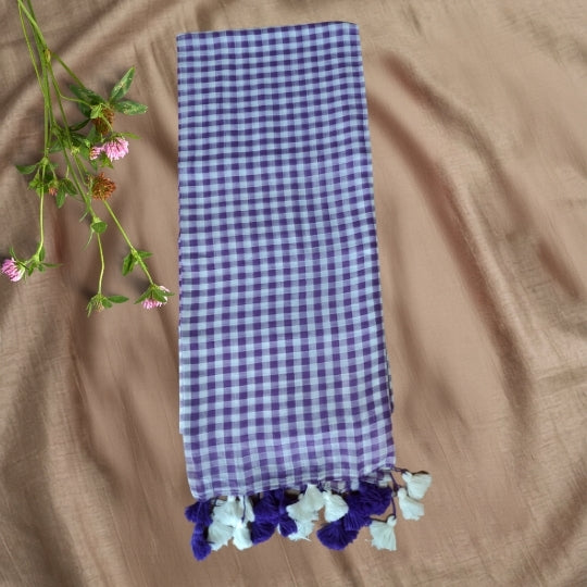 A purple and white gingham checkered scarf with decorative tassels, laid flat on a beige fabric background next to some small pink flowers.