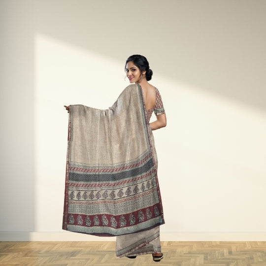 A woman models a light beige saree with a wide border featuring red, gray, and black patterns, standing against a minimalist beige wall with soft sunlight and a wooden floor.