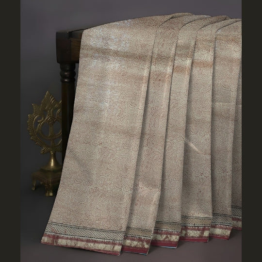 A draped beige and brown patterned fabric, likely a sari, with a decorative border, displayed over a dark wooden stand next to a brass decorative object against a dark gray background.
