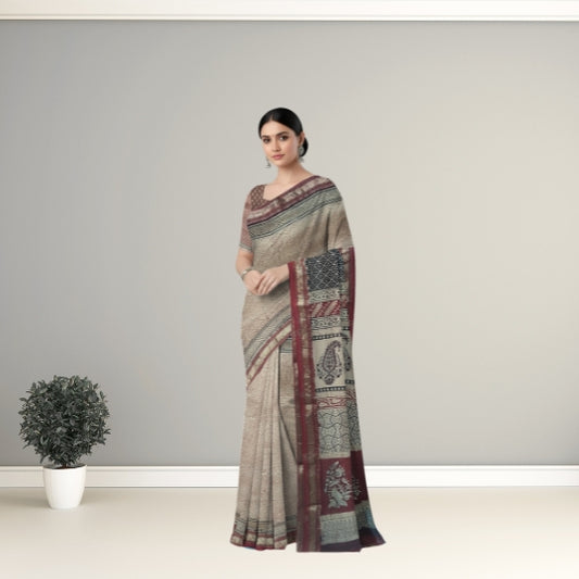 A woman modeling a beige and maroon patterned saree with a dark patterned blouse, standing in a minimalist studio setting with a small potted plant on the left.