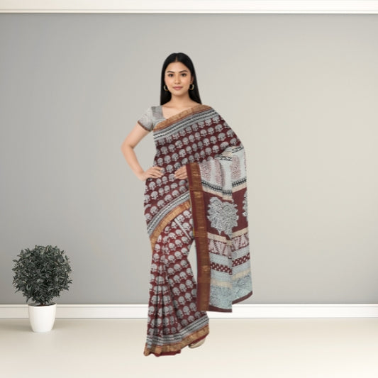 A woman modeling a printed saree with a matching blouse, standing against a plain gray wall with a small potted plant on the floor. The saree has a maroon and beige base with white floral patterns and a light blue patterned pallu.