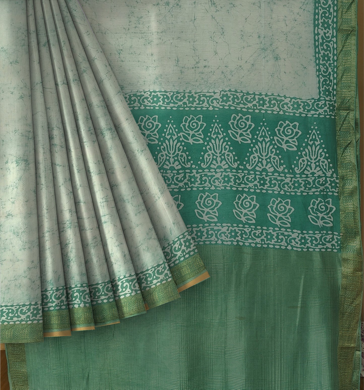 A close-up of a draped saree or fabric panel in sage green and white, featuring a wide border with white floral and geometric motifs and a gold-toned edge.