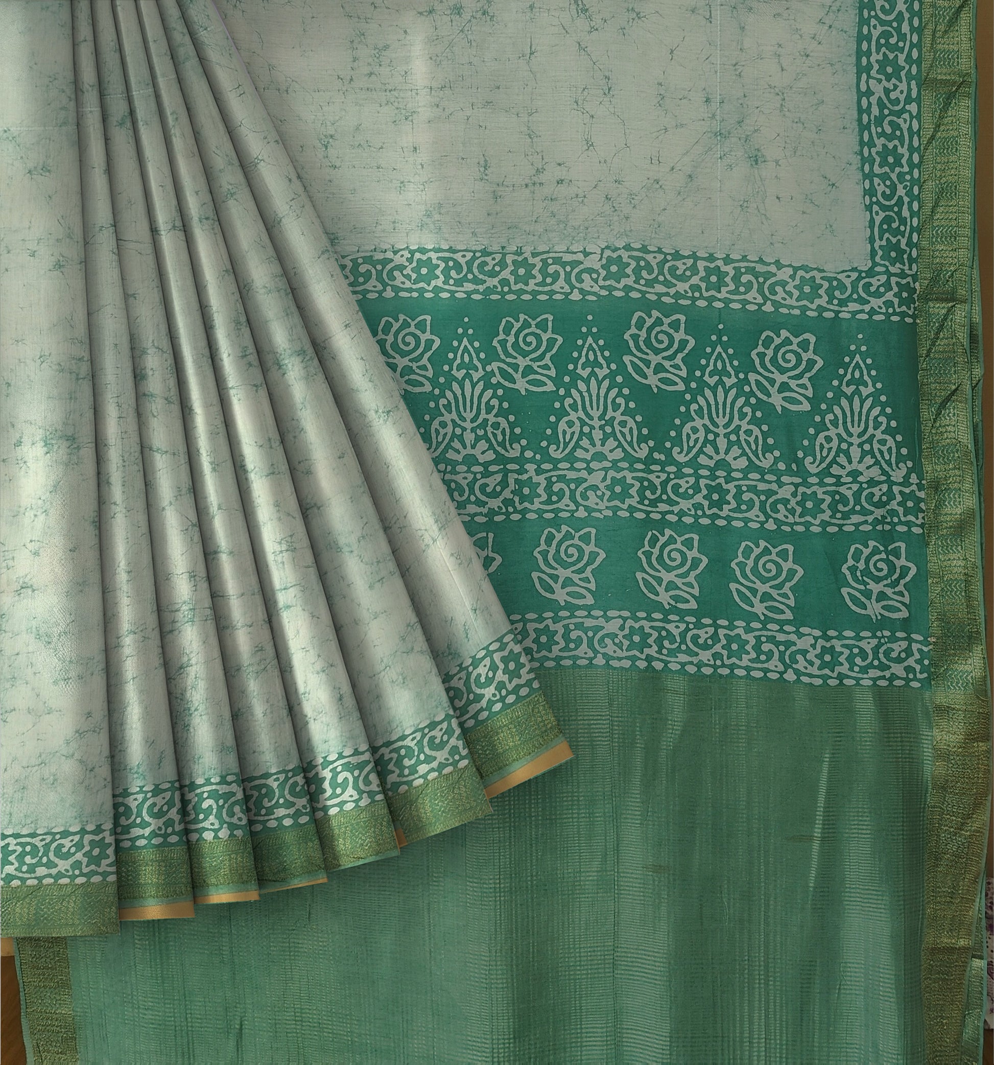 A close-up of a draped saree or fabric panel in sage green and white, featuring a wide border with white floral and geometric motifs and a gold-toned edge.