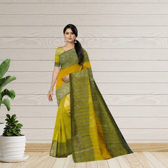 A woman modeling a yellow and olive green patterned saree with a matching blouse, standing against a light wood-paneled wall with a potted plant on the left.